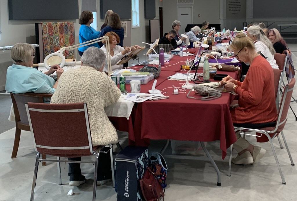 a large group of stitchers doing their stitching, some with magnifiers